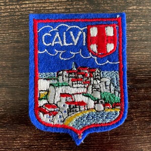 May include: Embroidered patch with a blue background and a red border. The patch features the word "CALVI" in white, a red and white shield, and a detailed illustration of a coastal town with red-roofed buildings. The patch is a souvenir.