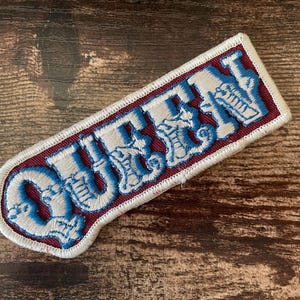 Peut inclure: Écusson brodé avec le mot "QUEEN" en blanc et bleu, souligné de blanc, sur un fond bordeaux. L'écusson a une bordure blanche et est rectangulaire avec des bords arrondis. L'écusson est sur une surface en bois.