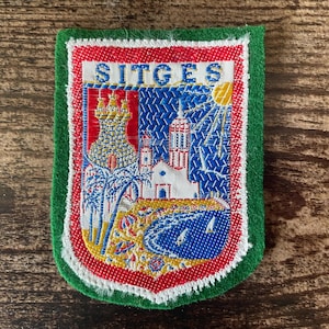 May include: A rectangular embroidered patch with a green felt border featuring the text SITGES at the top. The design depicts a coastal scene with a castle, a church, palm trees, a yellow sun, and a blue sea with small white sailboats.
