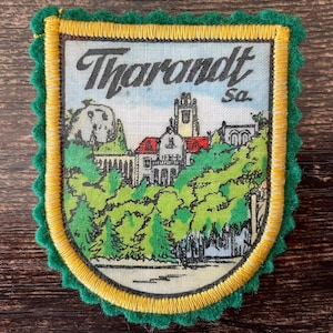 Pode incluir: Um emblema de tecido retangular com uma borda de feltro verde-escuro recortada e um contorno bordado em amarelo. O centro apresenta uma ilustração impressa de um complexo de edifícios com telhados vermelhos e árvores verdes, com o texto Tharandt sa. no topo.