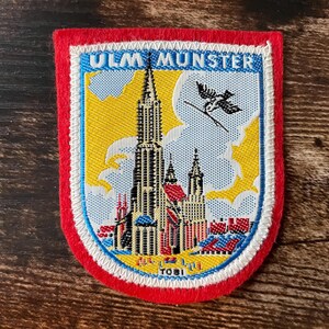 May include: A rectangular embroidered patch with a red border featuring the Ulm Minster cathedral in the center. The design includes a yellow sky, a black bird, and the text ULM MUNSTER at the top and TOBI at the bottom in blue lettering.