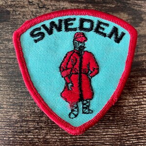 Pode incluir: Um emblema bordado em forma de escudo com fundo azul claro e borda vermelha. A palavra SWEDEN está bordada em letras maiúsculas pretas no topo. No centro, uma figura veste um casaco vermelho, um chapéu vermelho e faixas pretas nas pernas.