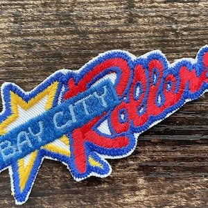 May include: An embroidered fabric patch featuring a yellow star background with a blue diagonal banner displaying the text BAY CITY in blue letters, overlaid with the word Rollers in red cursive script with a thick blue embroidered border.