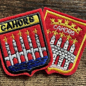 May include: Two shield-shaped embroidered patches featuring the text CAHORS. The designs display a red background with a white bridge, yellow fleur-de-lis accents, and blue water details. The patches have yellow and black stitched borders.