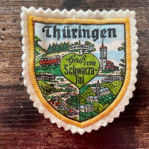 May include: A shield-shaped fabric patch with a scalloped cream-coloured felt border and yellow stitching. The centre features a colourful illustration of the Thüringen region, including a train, a tower, and a green heart with the text "Gruß vom Schwarza-Tal".