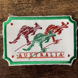 Peut inclure: Un écusson brodé rectangulaire avec un fond blanc et une bordure verte. Il présente deux silhouettes de kangourous rouges et une silhouette de kangourou verte au centre. Le mot AUSTRALIA est brodé en rouge sous les kangourous.