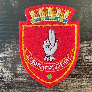 May include: Red embroidered patch with a shield shape, featuring a white hand gesture and the text "S. JEAN DE MAURIENNE". The patch has a yellow border and a crown design at the top.