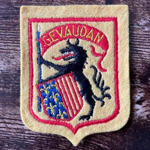 May include: A rectangular felt patch with a yellow background and a red border. It features an embroidered black wolf figure holding a blue shield with gold fleur-de-lis and red stripes. Above the wolf is a red banner with the text GEVAUDAN.