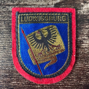 May include: A patch with a red border, black trim, and a blue center. The patch features the word "LUDWIGSBURG" in gold lettering at the top. The center design includes a gold and black eagle emblem with a red spear.