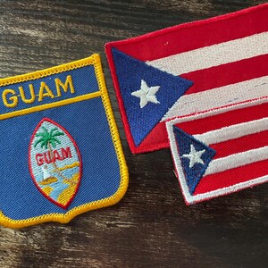 May include: Three embroidered patches: one blue with "GUAM" and a Guam seal, and two Puerto Rican flags. The Guam patch is shield-shaped with yellow trim. The Puerto Rican flags are red and white striped with a blue triangle and white star.