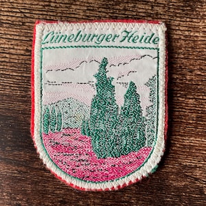 May include: Embroidered patch with a shield shape, featuring a landscape scene with green trees, pink ground, and a light pink sky. The text "Lüneburger Heide" is in green script at the top. The patch has a red border and white stitching.