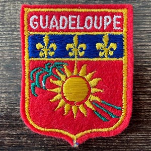 May include: A rectangular embroidered patch with a red background and a yellow border. The top section features the white text GUADELOUPE. Below is a blue horizontal band with three yellow fleur-de-lis, followed by a yellow sun and green palm fronds.