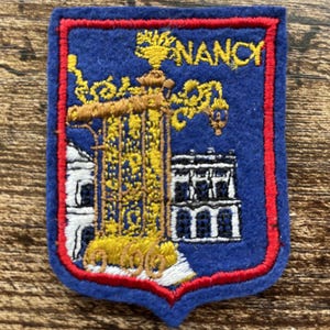 May include: A blue felt patch with a red border featuring an embroidered yellow ornate gate, white architectural details, and the word NANCY in yellow thread. The design depicts a landmark with intricate metalwork and a structured building facade.