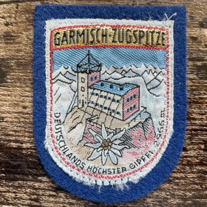 Könnte beinhalten: Ein blauer Filzaufnäher mit einer gestickten Berglandschaft, einem Gebäude auf einem Gipfel, einer weißen Edelweißblüte und dem Text GARMISCH-ZUGSPITZE sowie DEUTSCHLANDS HÖCHSTER GIPFEL 2965 m. Der Aufnäher hat einen weißen Rand und ein goldenes Banner.