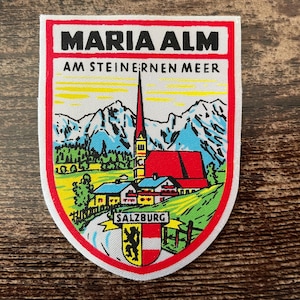 Könnte beinhalten: Schildförmiger Aufnäher mit dem Text "MARIA ALM AM STEINERNEN MEER" oben. Der Aufnäher zeigt eine farbenfrohe Illustration einer Kirche, Berge und das Wort "SALZBURG" unten, mit einem Wappen.