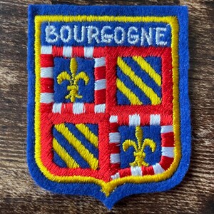 Pode incluir: Um emblema bordado retangular com uma borda de feltro azul apresentando o texto BOURGOGNE em branco. O design do escudo é dividido em quatro quadrantes com flores-de-lis amarelas e listras diagonais amarelas em campos azuis, emoldurados por bordas xadrez vermelhas e brancas e um contorno amarelo.