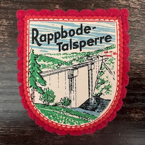 Peut inclure: Écusson souvenir vintage avec une bordure festonnée rouge. Le patch présente un paysage avec un pont et le texte "Rappbode-Talsperre" en noir. L'arrière-plan comprend un ciel bleu et des arbres verts.