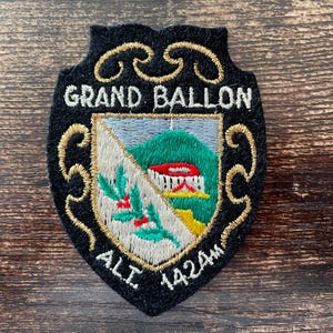 Könnte beinhalten: Ein schwarzer, schildförmiger Stickaufnäher mit dem Text GRAND BALLON und ALT. 1424M. Die Mitte zeigt ein Wappen mit einem grünen Berg, einem weißen Gebäude mit rotem Dach und einem Zweig mit grünen Blättern und roten Beeren auf weißem Grund.