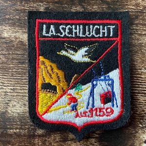 Può includere: Toppa ricamata con sfondo nero e bordo rosso. La toppa presenta il testo "LA.SCHLUCHT" in alto, un uccello in volo, una scena di montagna, uno sciatore e una seggiovia. Il numero "Auf 1159" è in basso.