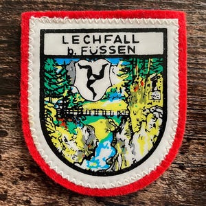 May include: A shield-shaped embroidered patch with a bright red border and white stitching. The center features a colorful graphic of the Lechfall waterfall near Fussen, including a bridge, trees, and a triskelion symbol with the text LECHFALL b. FUSSEN.