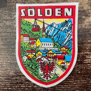 Könnte beinhalten: Ein schildförmiger Aufnäher mit dem Text SÖLDEN in Weiß auf rotem Grund. Das Design zeigt ein farbenfrohes Bergdorf mit Häusern, einer Kirche, einer Seilbahn und blauen Gipfeln. Ein rotes Adlerwappen ist im unteren Bereich mittig platziert.