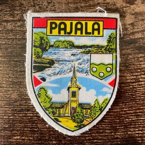 Peut inclure: Un écusson brodé en forme de bouclier portant le texte PAJALA dans un encadré jaune. Le motif représente une rivière bleue avec des rapides blancs, une église jaune avec un clocher, des arbres verts et un petit blason avec trois cercles.