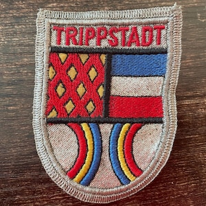 Puede incluir: Un parche bordado en forma de escudo con un borde plateado. La sección superior presenta el texto rojo TRIPPSTADT. Debajo, el lado izquierdo tiene un patrón de diamantes rojos sobre un fondo dorado, mientras que el lado derecho tiene rayas azules, blancas y rojas. Dos líneas curvas rojas, amarillas y azules están en la parte inferior.