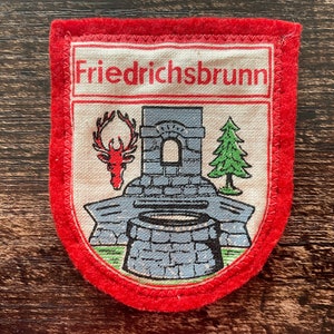 May include: A vintage patch with a red border and the word "Friedrichsbrunn" at the top. The patch features a deer head, a stone structure, and a green pine tree. The patch is likely a souvenir or emblem.