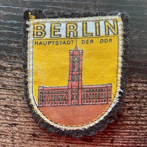 May include: A vintage Berlin patch with a yellow background and a red and brown illustration of a building. The patch has the word "BERLIN" at the top and "HAUPTSTADT DER DDR" below. The patch is outlined with a black border.