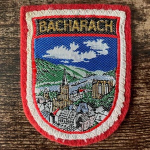 May include: A rectangular embroidered patch with a red border and a white stitched edge. The centre features a landscape of the town of Bacharach with a church, buildings, and green hills under a blue sky. The word BACHARACH is in gold text.