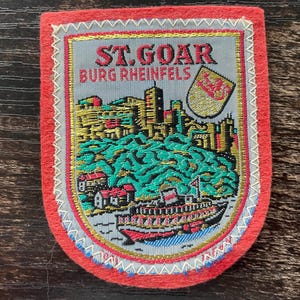 May include: A rectangular embroidered patch with a red felt border featuring a grey centre with the text ST. GOAR BURG RHEINFELS. The design depicts a castle on a hill, a green forest, a river, a boat, and a small shield with a red lion emblem.