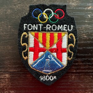Pode incluir: Um emblema bordado preto com os anéis olímpicos no topo, o texto FONT-ROMEU e um escudo central com listras verticais vermelhas e amarelas, uma cruz vermelha sobre branco e uma base azul com um ícone de torre branca, com o texto 1800M.
