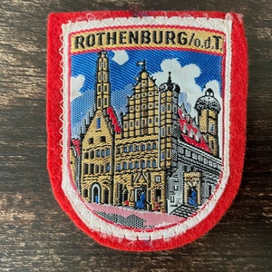 May include: A red and white embroidered patch with the text "ROTHENBURG/od.T." and a depiction of a medieval German town with a tall tower.
