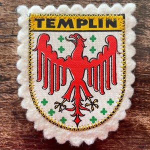 May include: A fabric patch with a scalloped white felt border featuring a shield design. The shield has a yellow top bar with the black text TEMPLIN, a white background with a red eagle, and small green cross symbols surrounding the eagle.