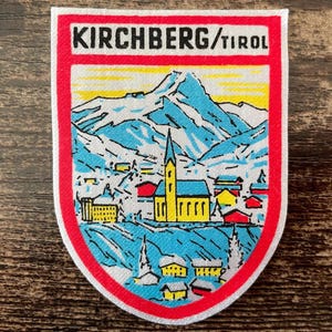 Op de afbeelding: Een rechthoekige stoffen patch met afgeronde onderste hoeken met een illustratie van een berglandschap. De tekst KIRCHBERG/TIROL staat in het zwart bovenaan. De scène toont een gele kerk, huizen en blauwe bergen.