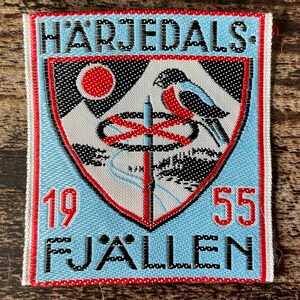 Può includere: Una toppa ricamata rettangolare con sfondo azzurro e bordo rosso. Presenta uno scudo con una montagna, un sole rosso, un uccello e un palo verticale. Il testo riporta HÄRJEDALS, 19 55 e FJÄLLEN in caratteri neri e rossi.