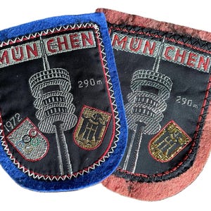 May include: Two embroidered patches featuring the Munich Olympic Tower with the text MUNCHEN and 290m. One patch has a blue felt border and the 1972 Olympic rings, while the other has a red felt border. Both display a black center with silver tower details.
