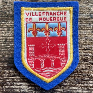Op de afbeelding: Een schildvormige geborduurde patch met een blauwe vilten rand. Het midden toont een wit wapenschild met de tekst VILLEFRANCHE DE ROUERGUE in het rood. Het bevat drie gouden fleur-de-lis, een rood veld met een wit kruis, twee torens en een brug.