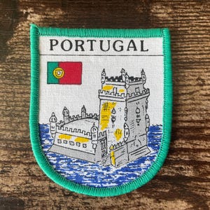 以下が含まれることがあります： 緑色の縁取りのある長方形のパッチには、「PORTUGAL」の文字と、ポルトガルの国旗と石造りの塔のイラストが描かれています。 塔はグレーで、黄色のアクセントがあり、青い水の背景に配置されています。