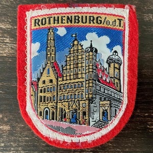 May include: A rectangular embroidered patch with a red felt border and a white stitched edge. The centre features a woven scene of a historic town with buildings, towers, and a blue sky with white clouds. The text ROTHENBURG/o.d.T. is at the top.
