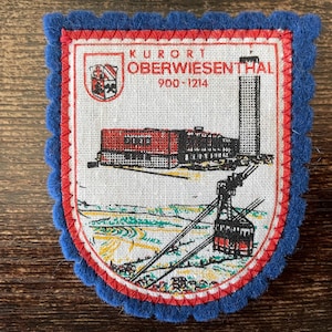 May include: A vintage patch with a blue scalloped border and a red and white inner border. The patch features a detailed illustration of a town with a cable car, buildings, and the text "KURORT OBERWIESENTHAL 900-1214".
