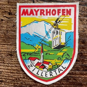 May include: A rectangular fabric patch with a red border featuring a mountain landscape, a cable car, and a village. The text MAYRHOFEN is printed in red at the top, and ZILLERTAL is printed in black on a white banner at the bottom of the design.