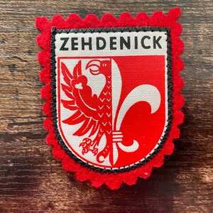 Peut inclure: Un écusson en tissu rectangulaire avec une bordure en feutrine rouge festonnée. Le centre présente un écu blanc avec un aigle rouge et une fleur de lys blanche. Le mot ZEHDENICK est imprimé en lettres capitales noires sur fond blanc.