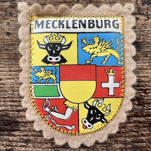 Op de afbeelding: Een rechthoekige patch met een geschulpte beige vilten rand met de tekst MECKLENBURG bovenaan. Het centrale ontwerp is een kleurrijk wapenschild verdeeld in secties met een zwarte stierenkop, een gele griffioen en een wit kruis.