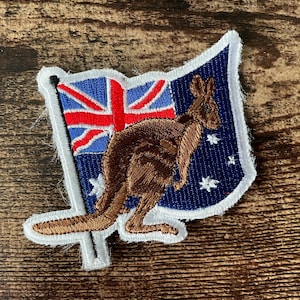 Peut inclure: Un écusson brodé représentant la silhouette d'un kangourou brun sur un drapeau bleu foncé avec l'Union Jack rouge et blanc dans le coin supérieur gauche et des étoiles blanches, le tout bordé d'une épaisse bordure blanche sur une base en tissu.