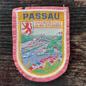 May include: A vintage Passau patch with a pink border. The patch features a colorful illustration of a city on a river, with the name "PASSAU" in blue letters at the top. The text "GRENZ/STADT an den 3 FLÜSSEN" is also visible.