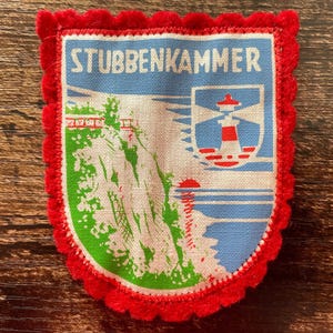 May include: A rectangular fabric patch with a scalloped red felt border. The centre features a printed illustration of a white cliff, a red and white lighthouse, and a red sun over blue water. The text STUBBENKAMMER is printed in white at the top.