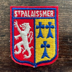May include: A rectangular embroidered patch featuring a shield design divided into two sections. The left side is red with a white lion, and the right side is yellow with three blue cross-like symbols. The text ST PALAIS SUR MER is in white.