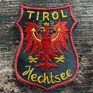 May include: A shield-shaped embroidered patch with a black background and a red border. It features a red eagle emblem with yellow accents, the text TIROL in yellow at the top, and the text Hechtsee in yellow cursive script at the bottom.