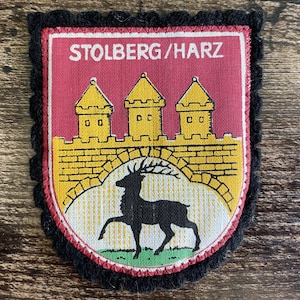 Könnte beinhalten: Ein schildförmiger Stoffaufnäher mit schwarzem, gewelltem Rand. Das Design zeigt einen roten Hintergrund mit dem weißen Schriftzug STOLBERG/HARZ, drei gelbe Türme über einer gelben Steinmauer und eine schwarze Hirschsilhouette auf weißem Grund.